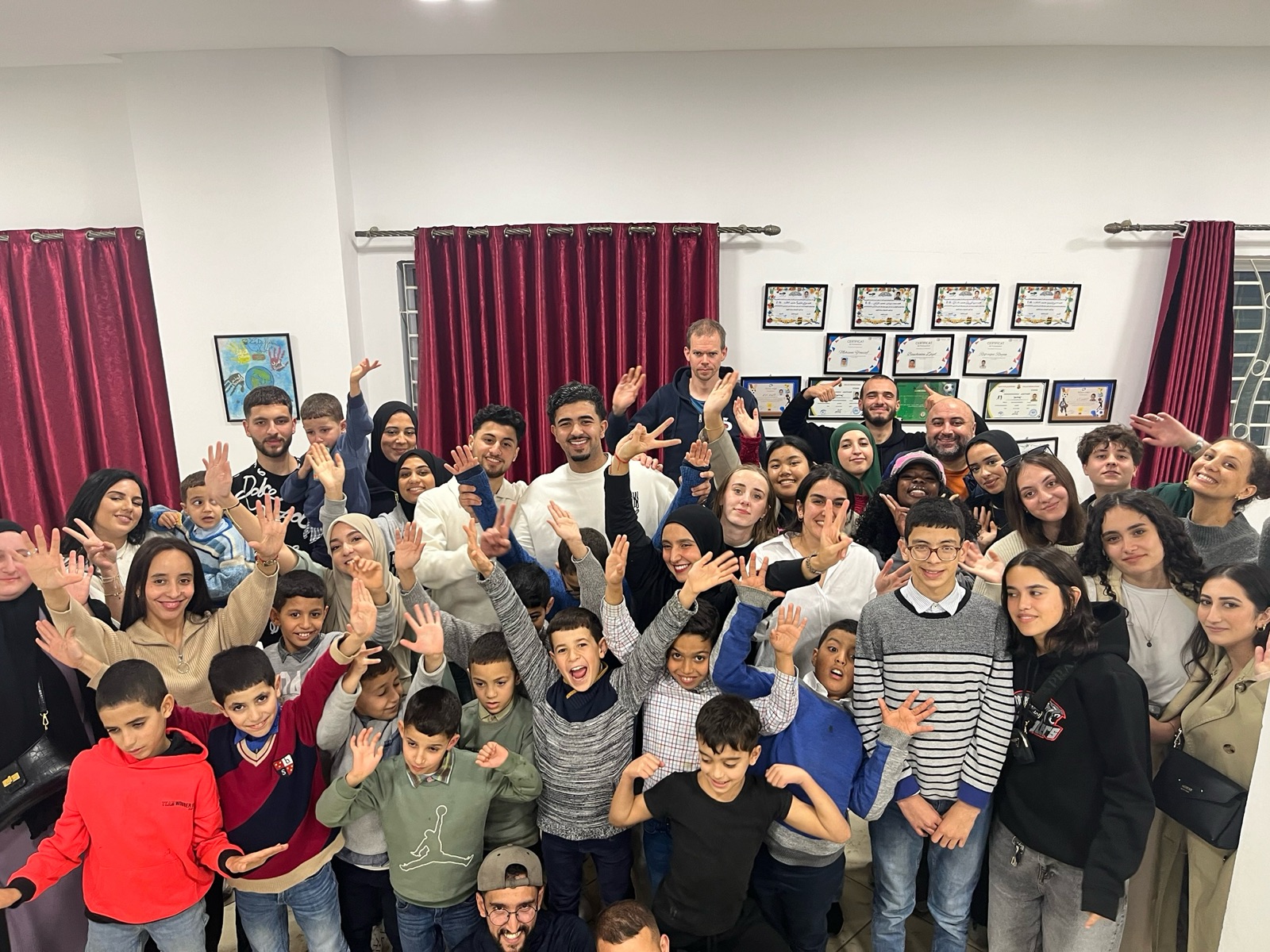 A diverse group of people posing together indoors, smiling and waving at the camera.