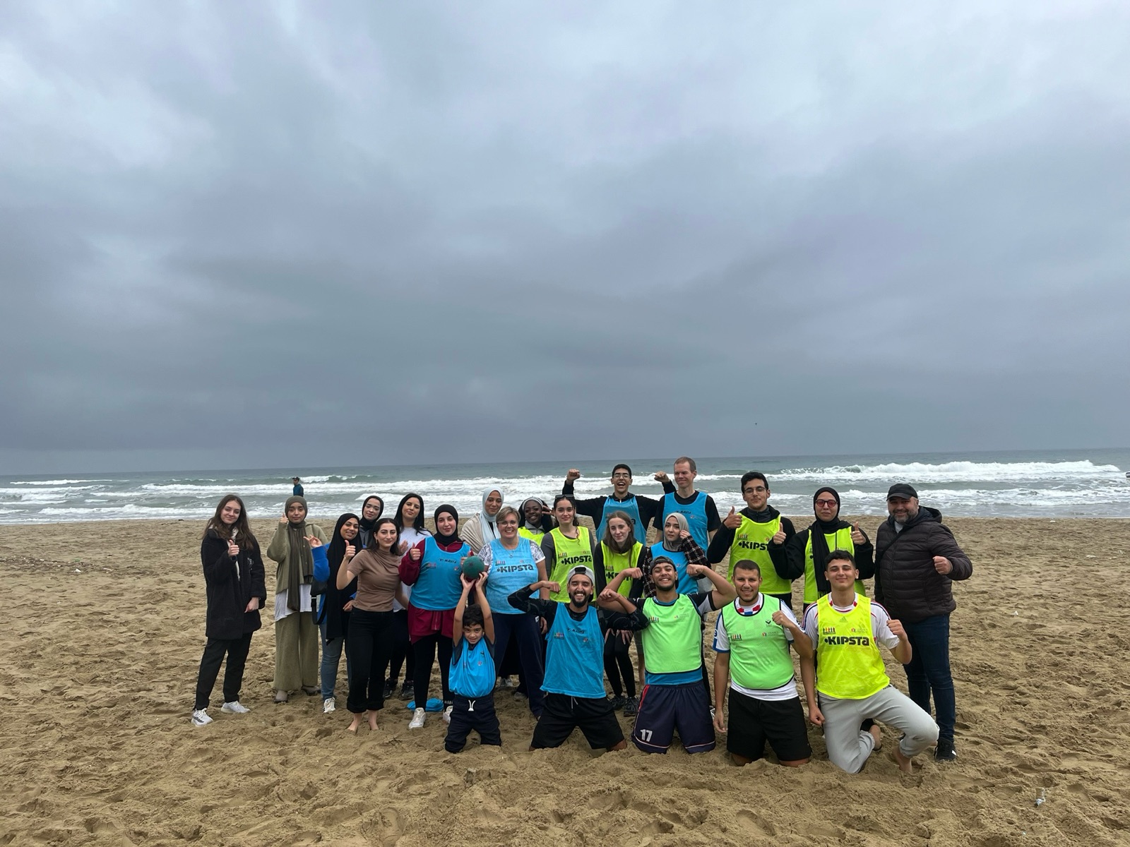 A group of people posing together on a sandy beach with the ocean and a cloudy sky in the background.
